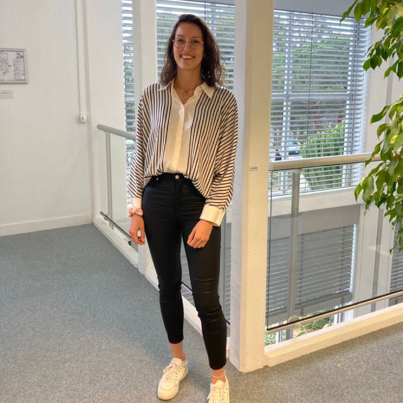 Une femme souriante se tenant devant un balcon avecu une plante venant de la droite